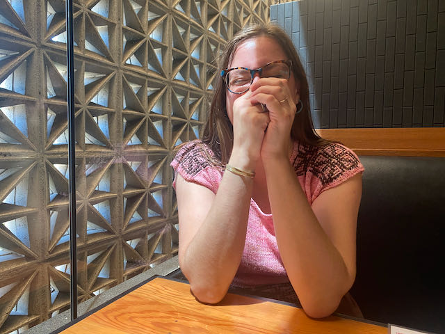 Allison, a white woman with brown hair and glasses, is laughing with her hands over her face. She wears a handknit sweater whose pattern echoes the breeze blocks behind her.