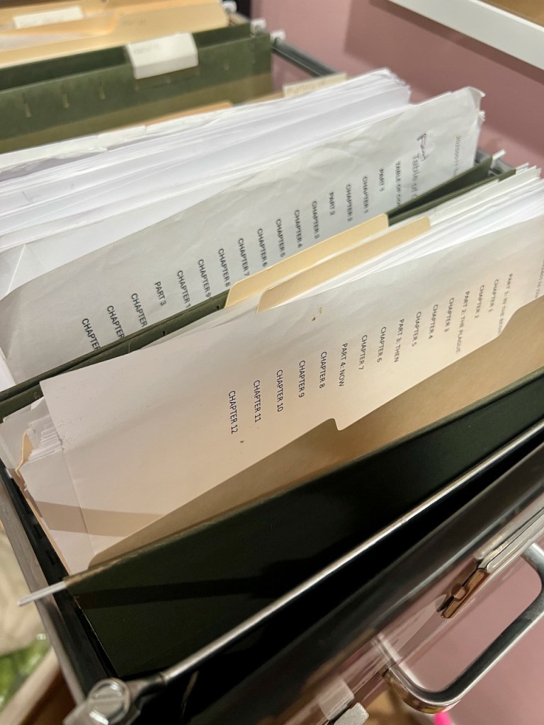 Printed drafts of a book in a filing cabinet drawer. Only the rumpled table of contents pages are visible inside the manila folders but both drafts are quite thick.