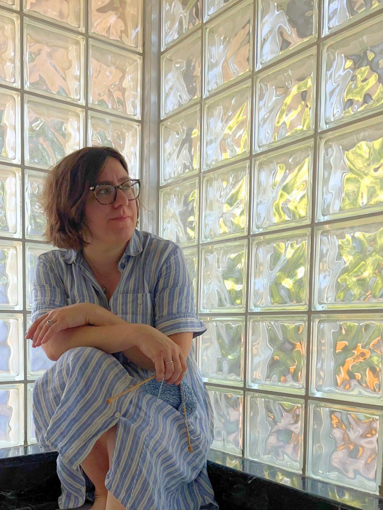 Allison, a white woman with brown hair and glasses, perches on a dark marble ledge in the corner between two glass block windows. She holds a small piece of knitting loosely and looks out the window to her left.